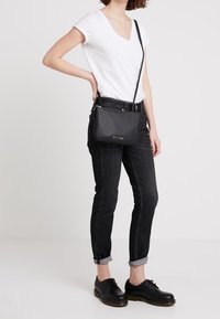 Czarna torebka na ramię z zamkiem błyskawicznym, gładka faktura skóry i subtelne oznaczenie marki. Połączona z ciemnymi, podwiniętymi dżinsami i czarnymi butami.