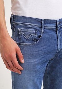 Jeans de mezclilla azul con un corte ajustado, que presentan un pequeño bolsillo trasero con un distintivo detalle de costura en forma de V y una textura suave.