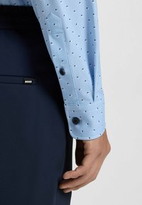 Camicia azzurro chiaro con un piccolo motivo geometrico, polsini abbottonati e pantaloni blu navy con un'etichetta di marca visibile. Tessuto liscio e vestibilità su misura.