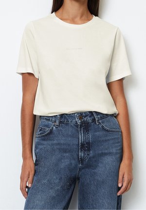 Mujer con una camiseta blanca de manga corta lisa metida en unos vaqueros azules de talle alto, sobre un fondo blanco.