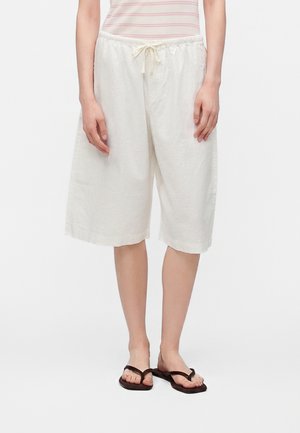 Personne portant un short blanc ample jusqu’aux genoux avec une taille à cordon, un haut rayé sans manches et des tongs noires, debout.
