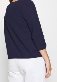 Mörkblå strukturerad topp med trekvartsärmar, med rundad halsringning och sidofickdetalj, ihop med vita byxor.