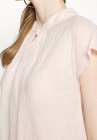 Blusa di lino rosa chiaro con una vestibilità rilassata, maniche corte, colletto rialzato e chiusura con un solo bottone al collo. Tessuto testurizzato.