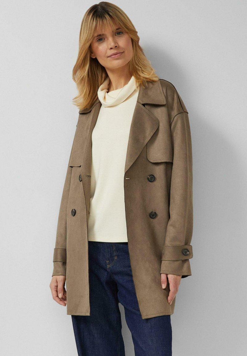 Blonde Frau in beigem, doppelt geknöpftem Wildledermantel über einem weißen Rollkragenpullover und dunklen blauen Jeans, stehend vor einem hellgrauen Hintergrund.