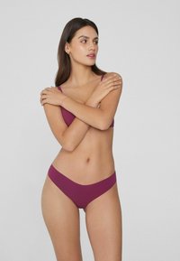 Femme portant un ensemble coordonné de soutien-gorge et de culotte violets, se tenant les bras croisés sur la poitrine, regardant sur le côté devant un fond gris.