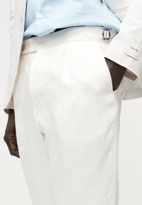 Personne portant un pantalon blanc sur mesure et une chemise bleu clair, avec une main dans la poche et une étiquette Calvin Klein visible sur la manche de la veste.