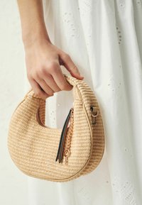 Main tenant un petit sac à main en paille tressée en forme de croissant avec une chaîne dorée et un pompon en cuir noir contre une robe blanche.