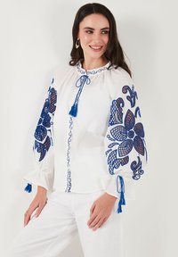 Blusa bianca con ricami blu che presentano motivi floreali. Maniche lunghe e imbottite con nappine e scollatura rotonda con cintura.