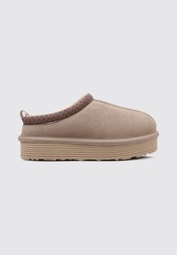 Zapato beige tipo slip-on con una superficie texturizada, ribete de patrón contrastante y suela estriada. Forma redondeada y diseño de perfil bajo.