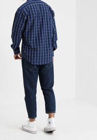 Chemise à carreaux bleue pour homme, en tissu coton, à manches longues, avec un ourlet arrondi. Portée avec un jean en denim noir, retroussé au niveau de la cheville, et des baskets blanches.