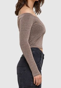 Glittrig brun långärmad topp med en av-axeln-design, figurnära passform och mjuk textur, tillsammans med högmidjade mörka jeans.