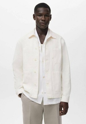 Uomo di colore che indossa una giacca cremisi con bottoni sopra una camicia bianca e pantaloni beige, in piedi con una mano nella tasca su uno sfondo semplice.