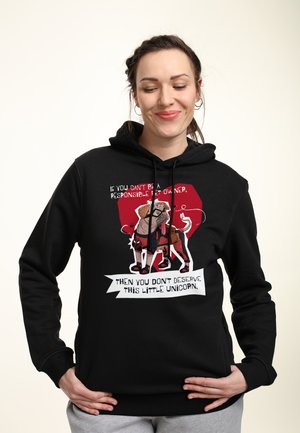 Kobieta w czarnej bluzie z kapturem z kreskówkowym mopsem przebraną za jednorożca oraz tekstem o odpowiedzialnym posiadaniu zwierząt, uśmiechająca się z zamkniętymi oczami.