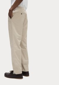 Beige Baumwoll-Chinos mit geradem Schnitt, seitlichen Eingrifftaschen und einer Gesäßtasche, die mit einem kleinen Metallknopf versehen ist, kombiniert mit dunklen Loafers.