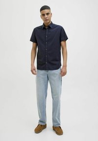 Camisa de botones de manga corta en color azul marino, de material de algodón, combinada con jeans de color azul claro y zapatos casuales marrones, de pie sobre un fondo neutro.