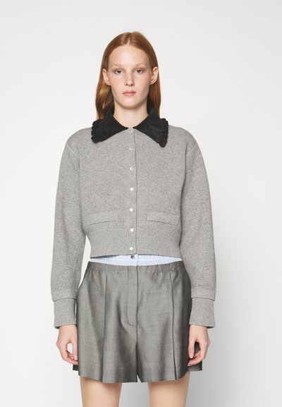 Pilkas trumpas megztinis su tekstūruotu juodu apykakliu, mygtukų priekyje ir dviem priekinėmis kišenėmis, derinamas su pilkais klostuotais šortais.