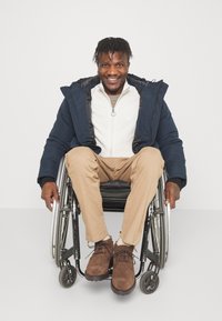 Tmavě modrá prošívaná bunda přes světle šedou zipovou mikinu, khaki kalhoty a hnědé semišové boty, sedící na vozíku na bílém pozadí.