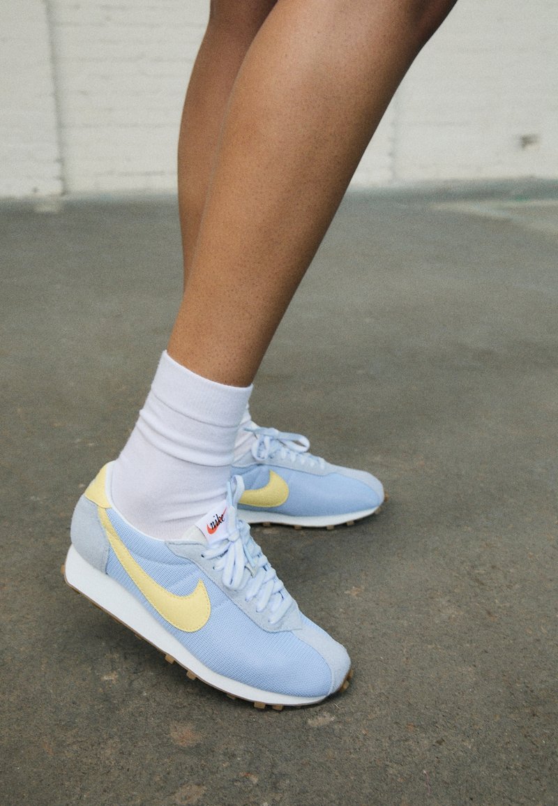 Jambes portant des chaussettes blanches et des baskets Nike bleu clair avec un swoosh jaune sur un sol en béton gris à l'intérieur.