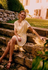 Jeune femme en robe jaune pâle et sandales, assise sur des marches en pierre en plein air, souriante, avec de la verdure et un bâtiment en arrière-plan.