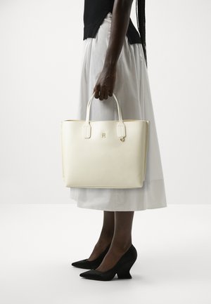 Borsa a tracolla in pelle crema con manici corti, accentuata da un logo dorato, di forma rettangolare. Il modello indossa una gonna bianca e tacchi a cuneo neri.