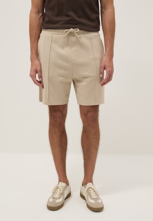 Homme portant un short beige à cordon et des baskets blanches, debout devant un fond clair uni.