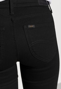 Vaqueros de mezclilla negra de corte ajustado, con un bolsillo trasero que presenta un sutil logo "Lee", costuras en contraste y una textura suave y mate.