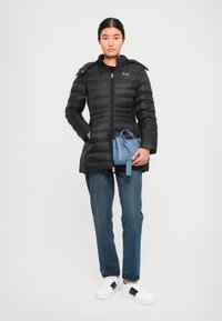 Zwarte pufferjack met een rits aan de voorkant, horizontale stiksels en gouden accenten. Gepaard met een blauwe handtas en blauwe jeans. Witte sneakers.