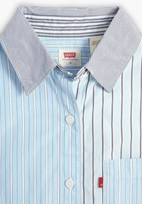 Camicia a righe azzurro chiaro con colletto a righe grigie in contrasto, bottoni bianchi e una piccola tasca con un'etichetta rossa.