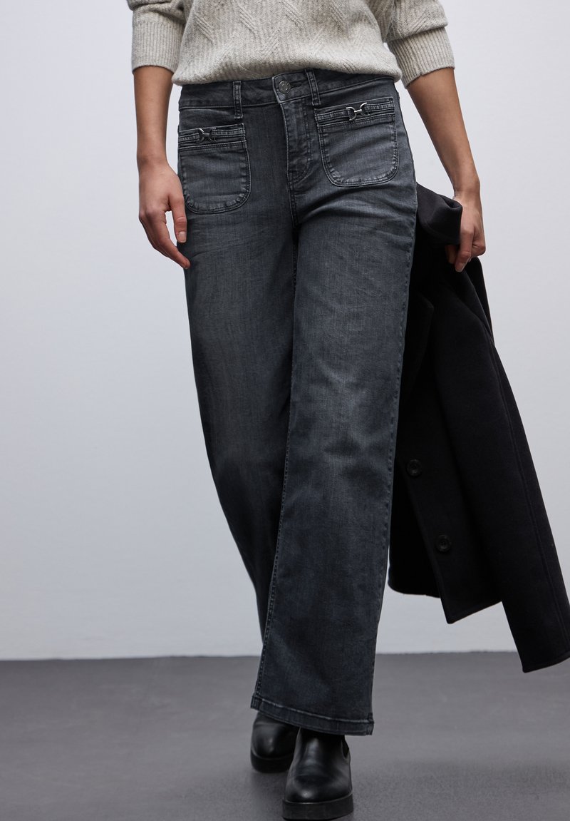 Jeans de pierna ancha en gris oscuro con bolsillos delanteros y una textura sutil, combinados con un suéter claro, sostenido con un abrigo negro en la mano.