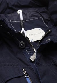 Intérieur d'une doudoune marine avec une fermeture éclair argentée, un cordon blanc et une étiquette avec le logo "bugatti". Tissu texturé avec des coutures contrastées.