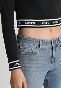 Svart kortärmad topp med långa ärmar, prydd med ett vitt band med "Levi's"-logotyp. Kombineras med ljusblå denimjeans som visar fickdetaljer.
