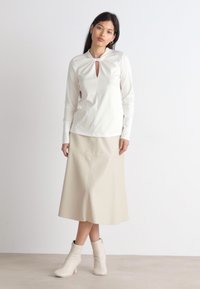 Hvit langermet bluse med nøkkelhull-detalj ved halsen, sammen med en beige A-linje skjørt og off-white ankelstøvler. Glatte teksturer.