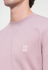 Svetloružová mikina s okrúhlym výstrihom a rebrovanými manžetami. Na prednej strane štvorcová nášivka s názvom značky "BOSS" v jemnej tónovej výšivke.