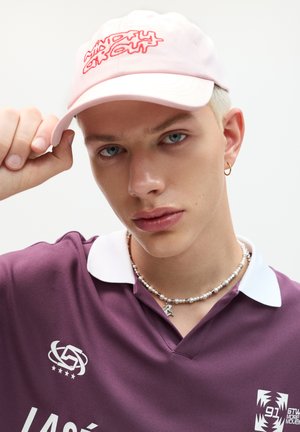 Casquette de baseball rose clair avec du texte rouge brodé, portée avec un polo violet sombre doté d'un col blanc et d'un collier de perles.
