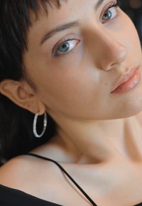 Close-up de uma pessoa com pele clara, vestindo uma blusa preta e brincos de argola prateados brilhantes, apresentando pele suave e cor de batom neutra.