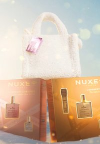 Vit fluffig handväska med två NUXE produktkartonger. Gyllene och rosa förpackning med tydlig märkning, produktnamn och ett transparent fönster.