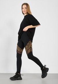 T-shirt oversize nero abbinato a leggings neri con stampa leopardata e pannelli in rete, completato da scarpe da ginnastica nere.