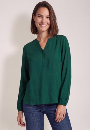 Femme souriante aux cheveux bruns mi-longs, vêtue d'un chemisier vert à manches longues et d'un jean bleu sur un fond uni.