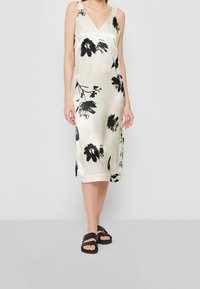 Satinová šaty na ramínka v krémové barvě s černým květinovým potiskem, s V-výstřihem a širokými ramínky; spárované s černými šlapkami. Hladká textura.