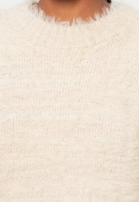 Maglione crema con texture, dalla superficie morbida e pelosa, presenta un collo a coste e una vestibilità leggermente ampia per maggiore comodità.