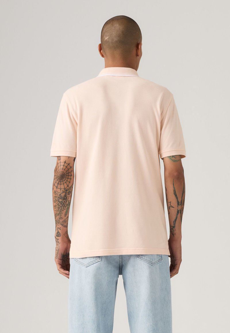 Homme avec des tatouages sur les bras, portant un polo rose pâle à manches courtes et un jean bleu clair, debout de dos sur un fond uni.