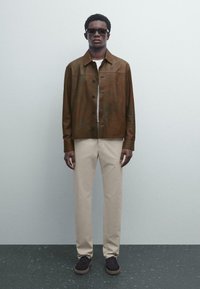 Man wearing brown leather jacket, white shirt, beige trousers, black shoes, and dark sunglasses standing against a light gray wall.
