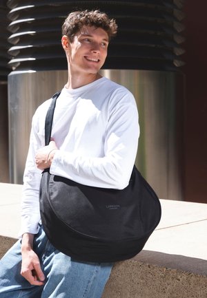 Svart axelväska i texturerat tyg, med oval form och ett tryckt logotyp. Bärs som crossbody mot en ljus bakgrund.