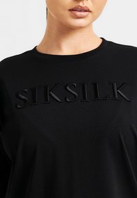 Černé tričko s výrazným černým vyšitým logem "SIKSILK" na přední straně, vyrobené z měkké bavlněné tkaniny s kulatým výstřihem.