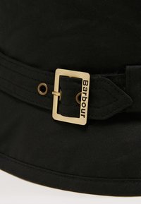 Un chapeau seau noir en tissu avec une finition texturée, featuring une boucle en métal doré avec le nom de la marque "Barbour" en relief.
