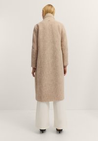 Langer, beigefarbener, strukturierter Mantel mit hohem Kragen und langen Ärmeln, der eine lockere Passform und minimale Taschenstiche auf der Rückseite aufweist.
