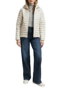 Lichtbeige pufferjack met capuchon, doorgestikte textuur, voorrits, gedragen over een grijze coltrui en donkerblauwe wijde jeans.