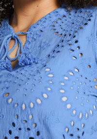 Blusa azzurra chiara con motivi floreali ricamati e occhiaie. I lacci al collo con frange aggiungono texture e dettaglio.