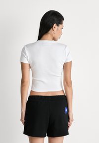 Valkoinen, lyhythihainen, ribbikuvioitu crop-top, jonka parina mustat shortsit, joissa on sininen kukkakuosi vasemmassa lahkeessa. Sujuva materiaali, istuva siluetti.