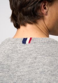 Grå stickad tröja med ribbad krage och en dekorativ trefärgad patch i blått, vitt och rött på baksidan.